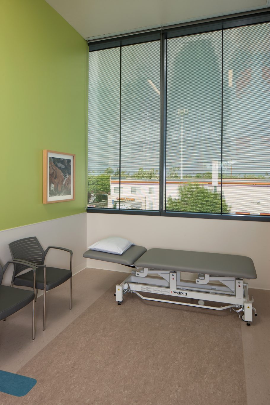 Shriners Childrens Hospital, Pasadena, US Clinical room with a treatment table, chair, and large windows overlooking greenery.