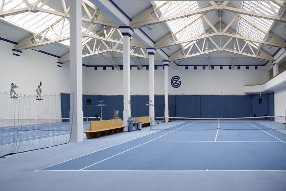 Indoor tennis court with blue surfaces and high ceilings, featuring a seating area.